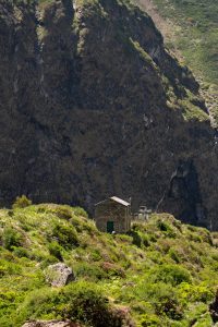 Small stone hut with a metal roof on a green shrub-covered slope beneath a large dark cliff