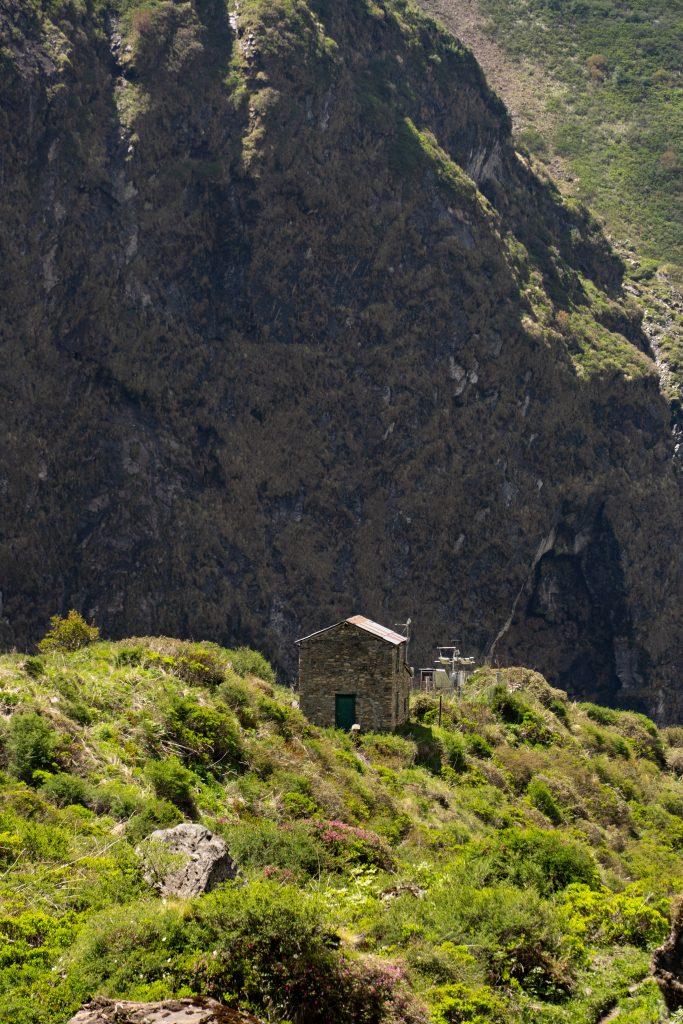 Small stone hut with a metal roof on a green shrub-covered slope beneath a large dark cliff