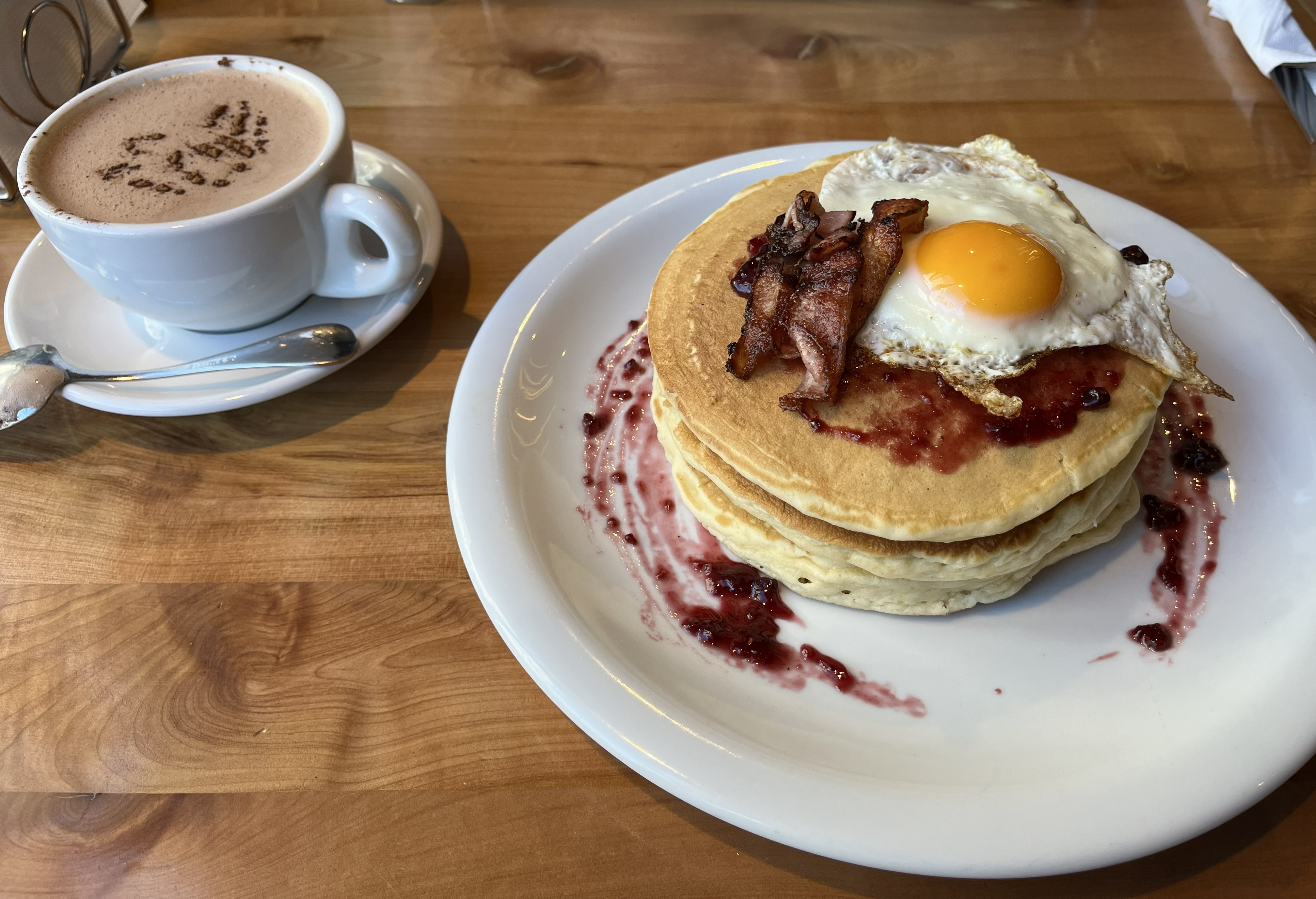 A plate of three fluffy pancakes stacked on top of each other, topped with a fried egg that has a bright yellow yolk. Strips of crispy bacon are placed on top of the egg, and there appears to be a drizzle of berry jam around the pancakes.