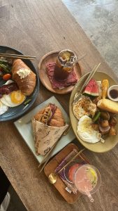 Breakfast spread on a wooden table with eggs, bacon, green beans, fried plantains, fresh fruit, a bacon croissant sandwich, and a pink iced drink with lime, served with gold utensils in a cozy rustic setting.