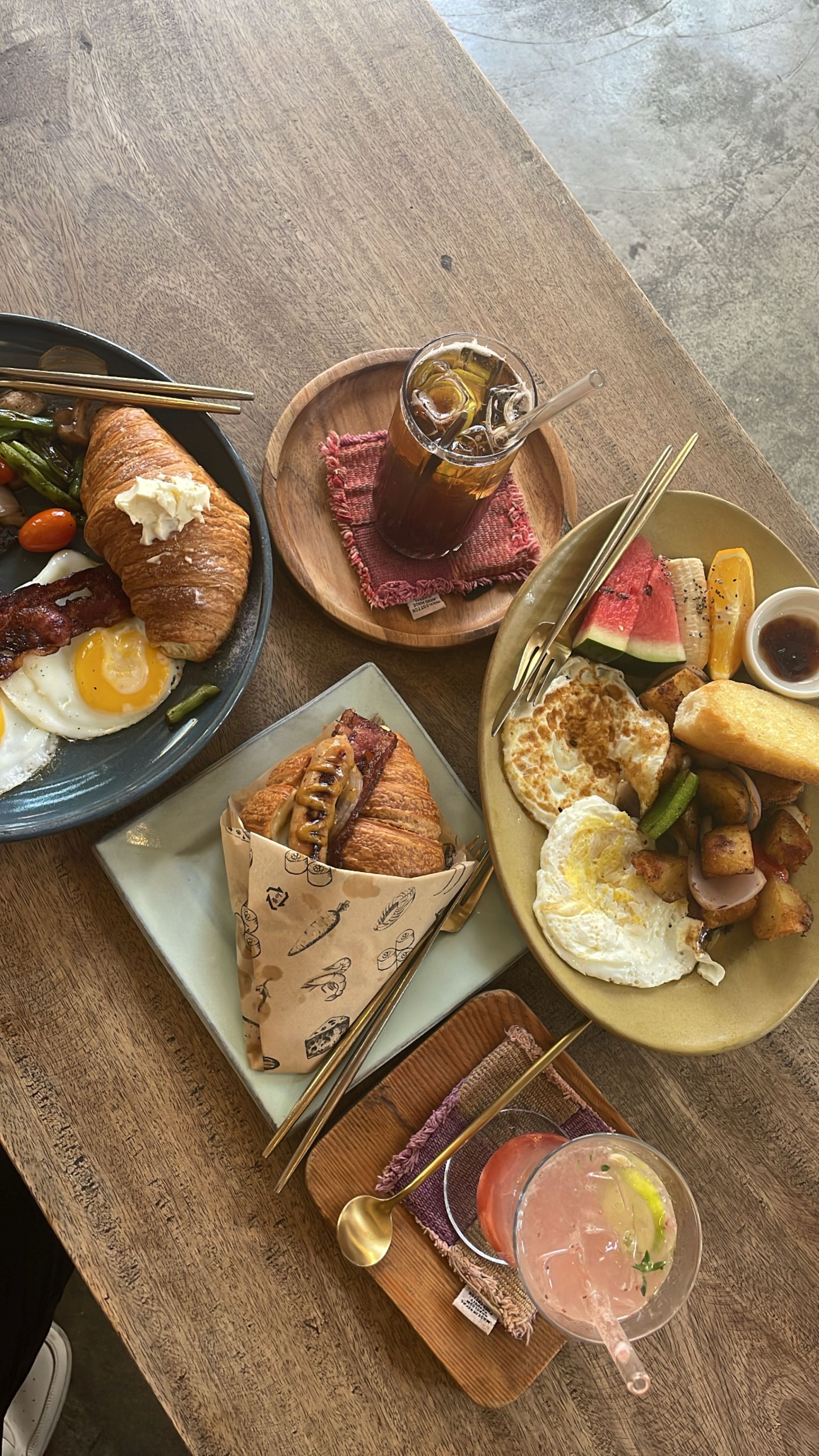 Breakfast spread on a wooden table with eggs, bacon, green beans, fried plantains, fresh fruit, a bacon croissant sandwich, and a pink iced drink with lime, served with gold utensils in a cozy rustic setting.