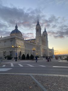 Almudena Cathedral at sunset in Madrid, Spain, with illuminated street lamps and a cloudy sky.
