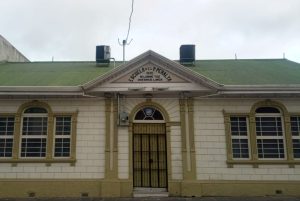 Facade of the Escuela del Padre Peralta (1845) in Cartago, Costa Rica. Cream-colored building with green corrugated roof, a pediment bearing the school's name and Latin motto, arched windows and a black wrought-iron entrance gate.