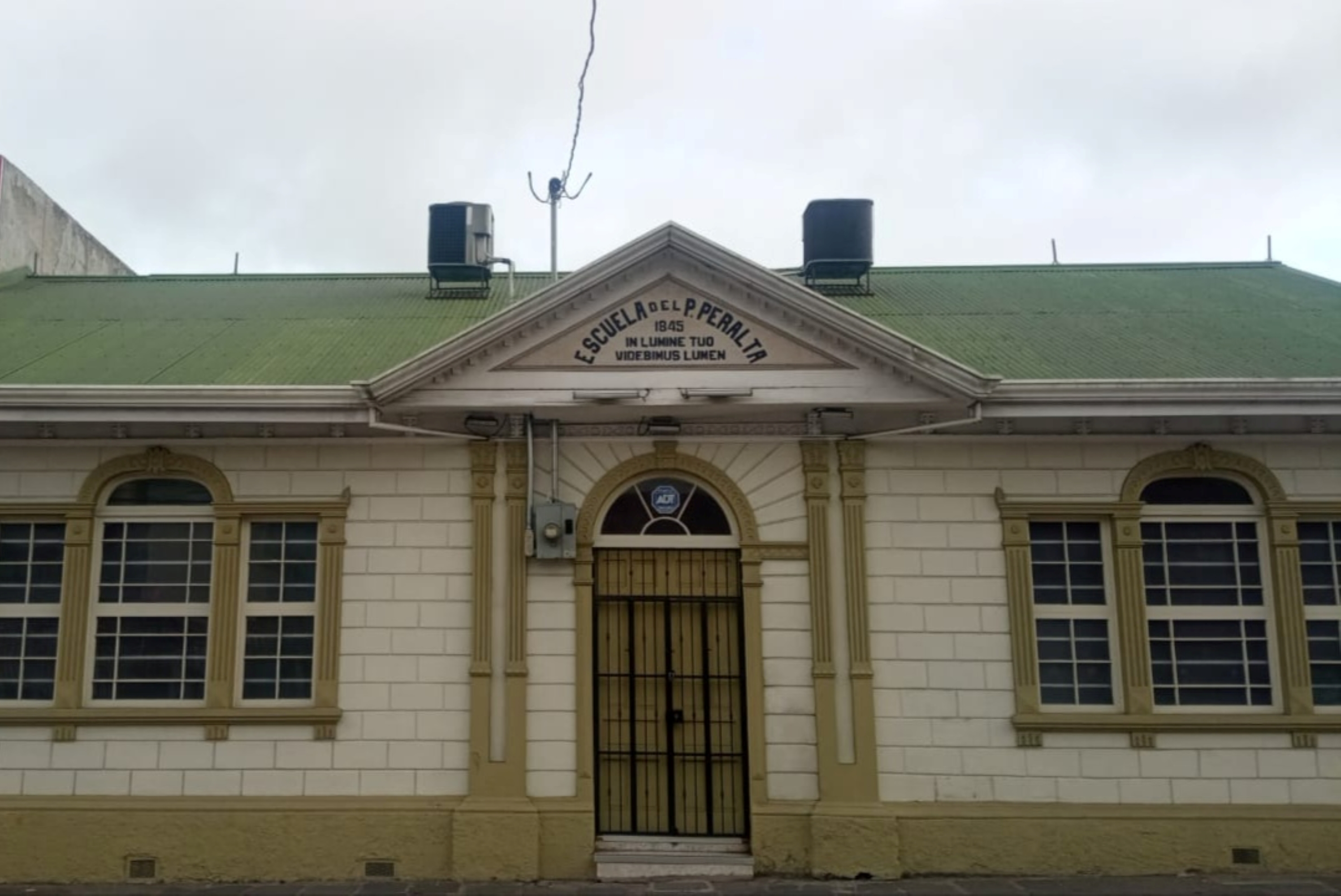 Facade of the Escuela del Padre Peralta (1845) in Cartago, Costa Rica. Cream-colored building with green corrugated roof, a pediment bearing the school's name and Latin motto, arched windows and a black wrought-iron entrance gate.