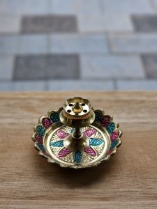 A small handcrafted brass incense stick holder with colorful floral designs sits on a wooden surface. The shallow depth of field draws attention to its intricate details and traditional craftsmanship. Captured in Pantheeramkavu, Kozhikode, Kerala.