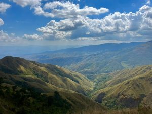 Paisaje montañoso con colinas, un valle y un cielo azul con nubes blancas.