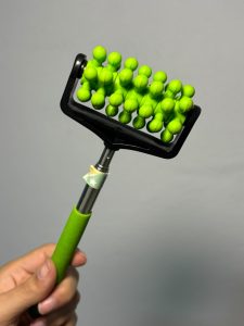 Close-up photograph of a green handheld massage roller with multiple rounded massage nodes, held in a person's hand against a plain gray background.