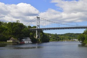 Suspension bridge spanning a calm river, with a paddlewheel boat docked below and green trees lining both riverbanks under a cloudy sky