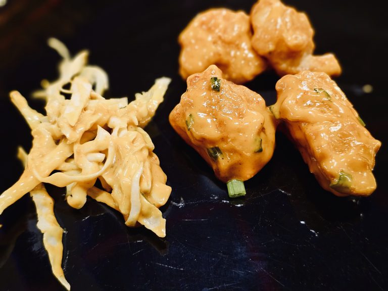 Crispy bite-sized chicken dynamite, coated in a creamy orange sauce, is served with a fresh cabbage salad on a black plate. The contrasting colors and textures make the dish look delicious. Captured in Mankavu, Kozhikode.