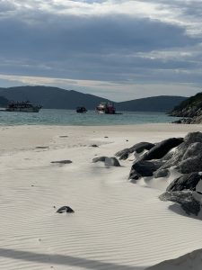White sandy beach with ripples, calm turquoise water, boats, and distant mountains under a cloudy sky.