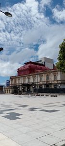 Rear view of the National Theater of Costa Rica in San José, showing its red domed roof and neoclassical stone facade rising above a modern plaza, with a high-rise building in the background.