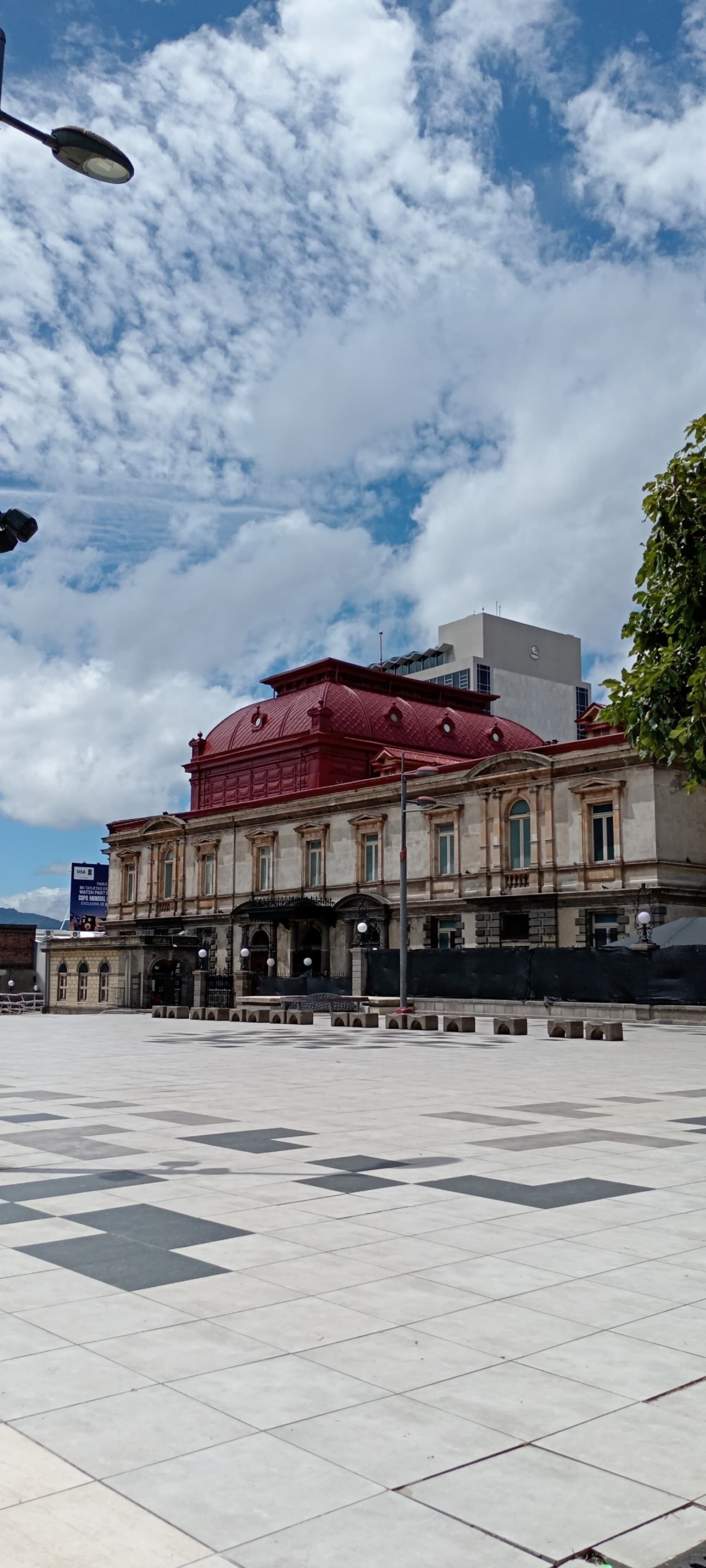 Rear view of the National Theater of Costa Rica in San José, showing its red domed roof and neoclassical stone facade rising above a modern plaza, with a high-rise building in the background.