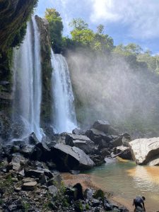 Waterfall flowing over rocks with mist, sunlight, and lush green vegetation.