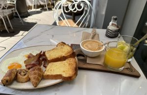 A breakfast spread is displayed on a marble table. On one side of the plate, there are two sunny-side-up eggs with bright yellow yolks, crispy bacon strips, and two slices of thick, toasted bread.On the other side of the table, there is a wooden serving board that holds a bowl of creamy coffee with a swirled surface, along with two glasses.