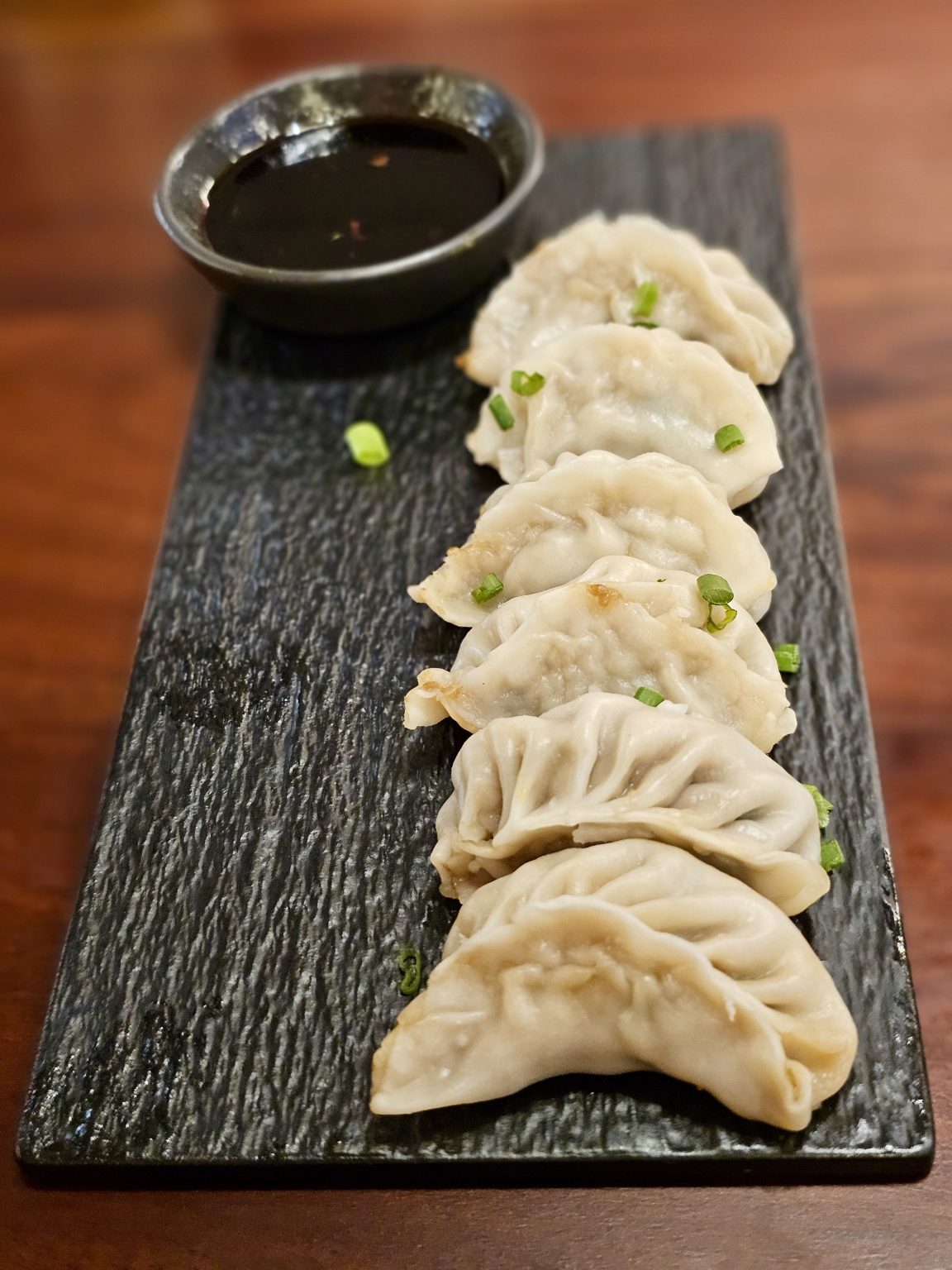 A row of freshly steamed chicken dumplings (momos) is served on a black stone platter with a bowl of dipping sauce. The simple presentation and soft texture make this dish look warm and inviting. Captured in Mankavu, Kozhikode.
