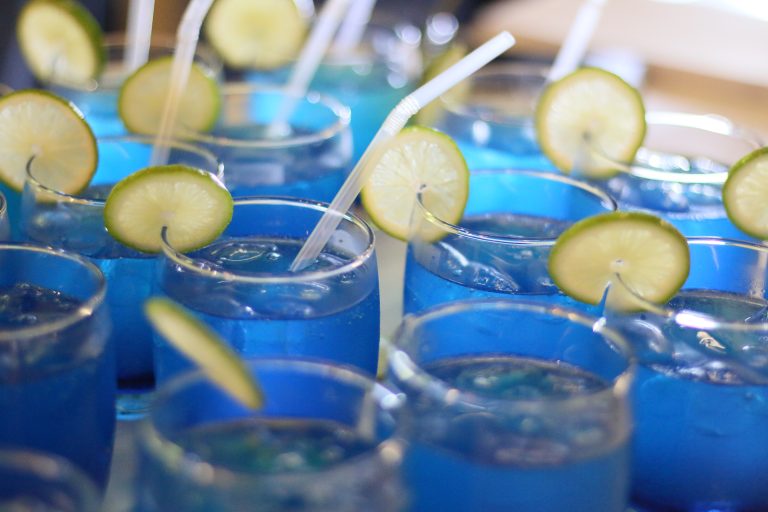A vibrant array of bright blue mocktails filled with ice, each served in a stemless glass. They are garnished with fresh lime slices and white straws, creating a refreshing, tropical party vibe.