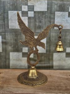A decorative brass eagle holds a small hanging bell, showcasing fine traditional metalwork. The blurred background keeps the focus on the detailed craftsmanship. Captured in Pantheeramkavu, Kozhikode, Kerala.