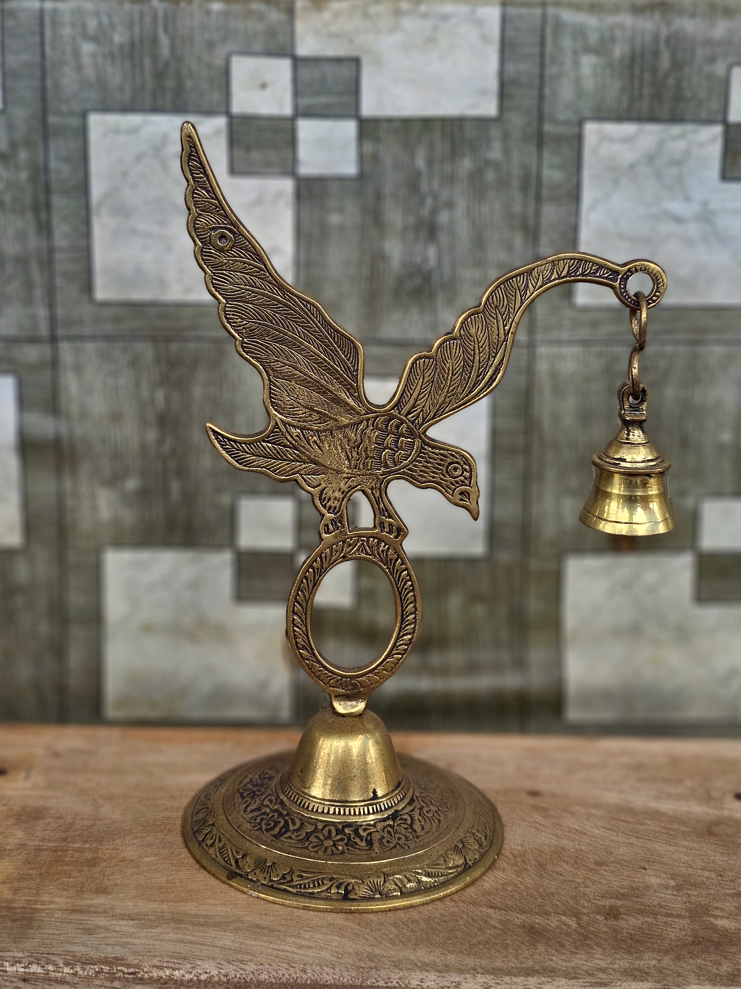 A decorative brass eagle holds a small hanging bell, showcasing fine traditional metalwork. The blurred background keeps the focus on the detailed craftsmanship. Captured in Pantheeramkavu, Kozhikode, Kerala.