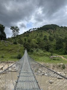 A metal suspension footbridge with chain-link mesh sides stretches forward over a wide, muddy riverbed towards green, terraced hillsides. Lush trees cover the landscape, and a tall solitary tree stands on a grassy slope to the left. In the background, forested green hills rise under a dark, overcast, and cloudy sky.
