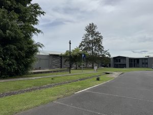 Wide exterior view of modern institutional buildings surrounded by green lawns, mature trees, paved walkways, a drainage channel, lampposts, and a curved asphalt road under a cloudy sky.