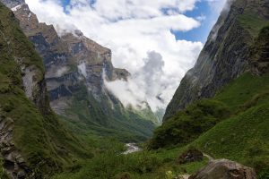 A lush, green valley stretches between towering, rocky mountains. A river winds through the landscape, with fluffy clouds and a blue sky overhead.
