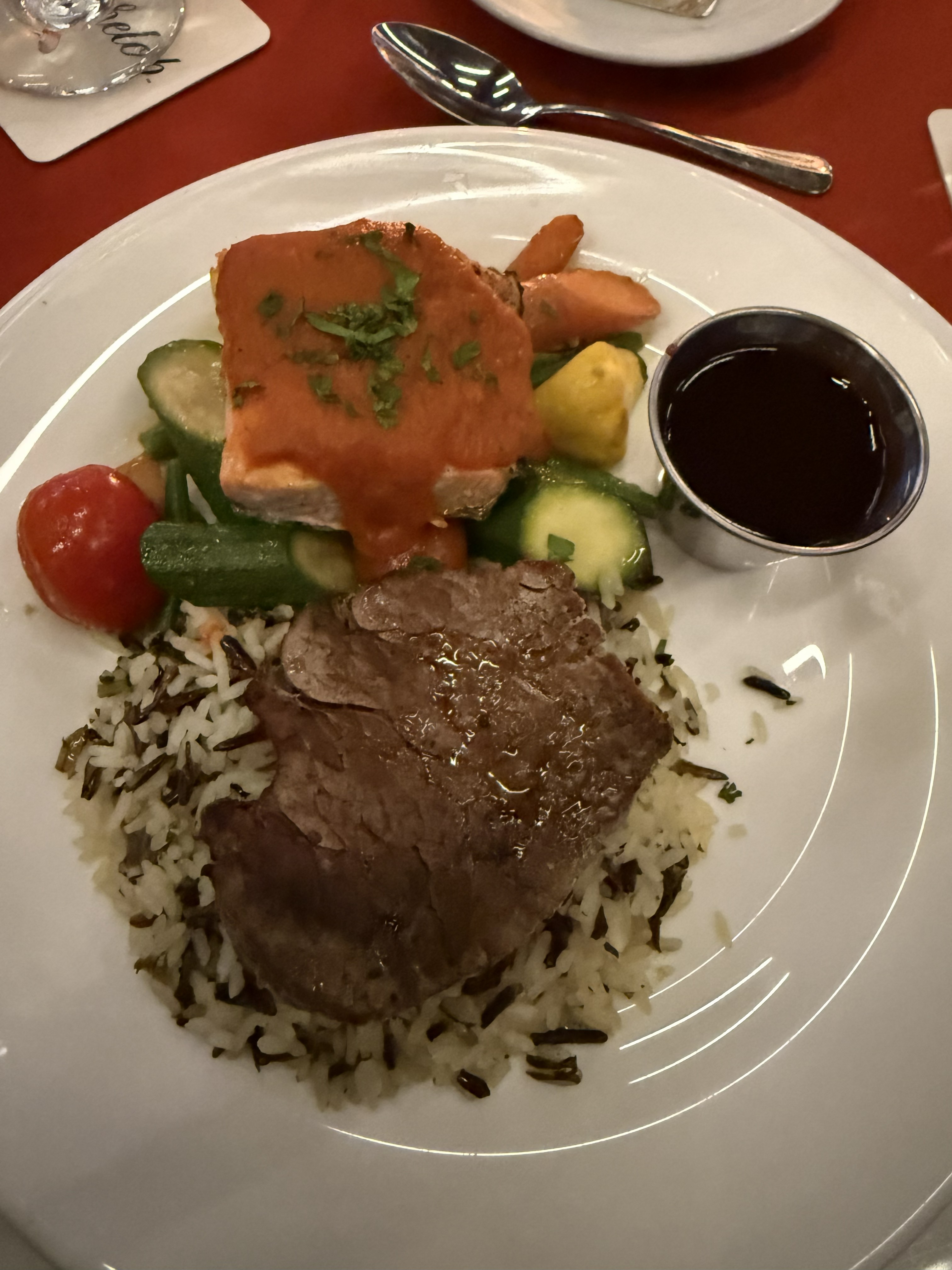 On top of the rice, there is a serving of tender, sliced beef, glistening with a savory sheen. Above the beef, a colorful assortment of vegetables includes vibrant orange carrots, green zucchini, and crisp green beans. A piece of salmon sits prominently atop the vegetables, garnished with a rich, reddish-orange sauce and a sprinkle of fresh herbs.