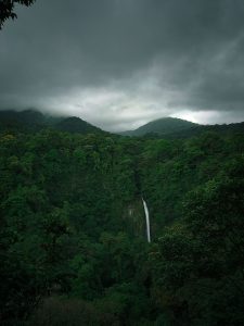 A tall waterfall drops through a dense green forest, surrounded by misty hills beneath a dark cloudy sky.