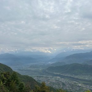 Panoramic view of green hills, a winding river, and snow-capped mountains beneath a cloudy sky.