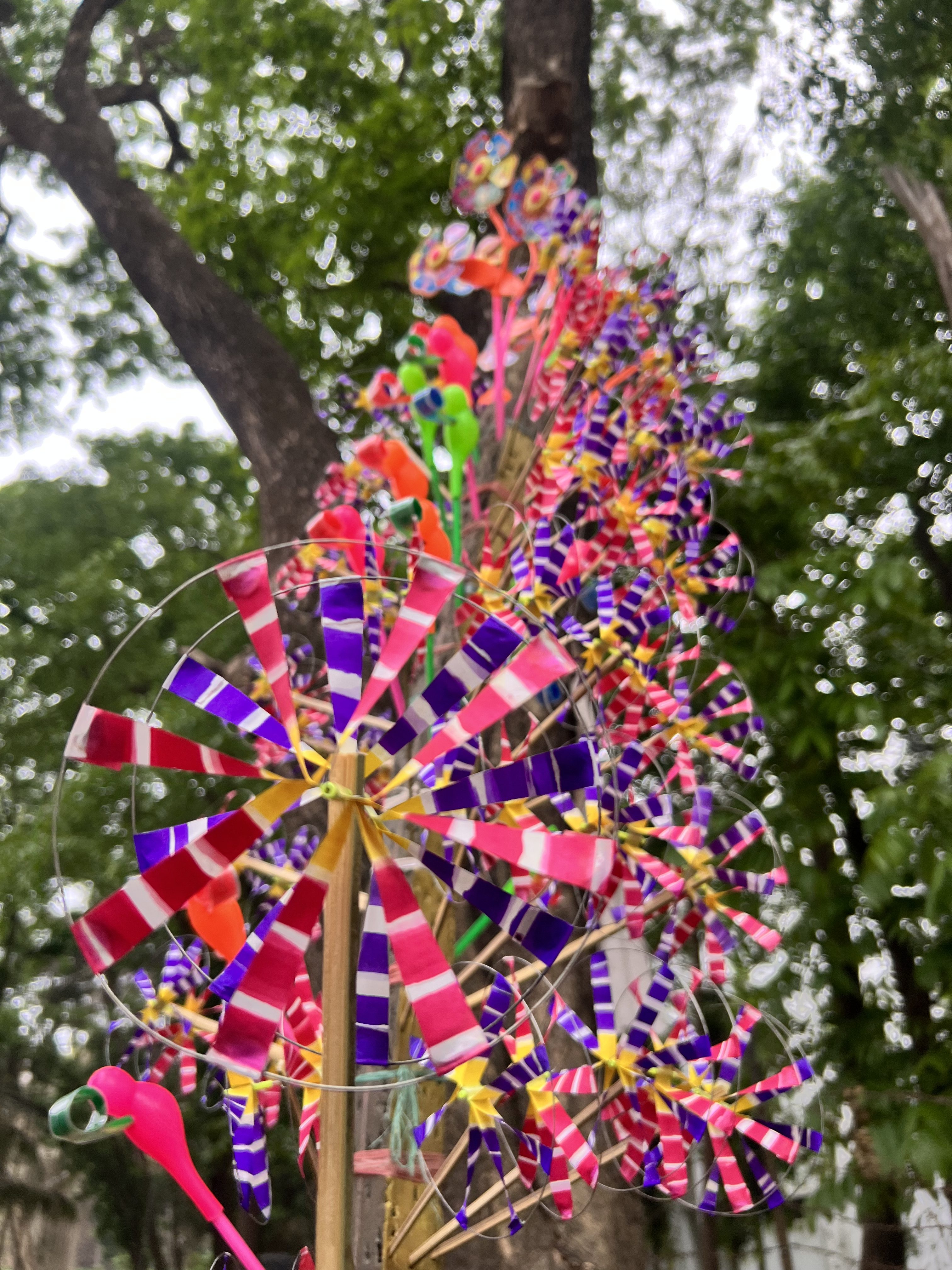 A close-up view of a vibrant, multi-colored paper pinwheel spinning toy, part of a larger cluster mounted on a bamboo stick with green trees blurred in the background.