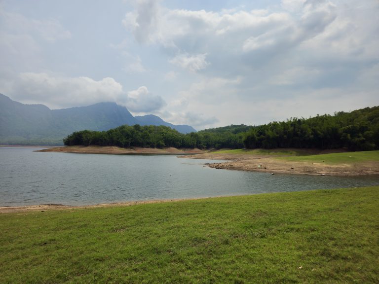 A peaceful reservoir surrounded by lush green forests and mist-covered hills under a dramatic cloudy sky. The calm water and open landscape create a beautiful view of nature. Captured in Parambikulam Tiger Reserve, Palakkad, Kerala.