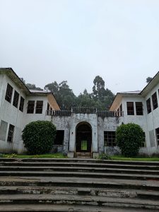Symmetrical facade of a former patient pavilion at the Sanatorio Durán in Costa Rica, with a central arched entrance flanked by twin wings and dense trees behind.
