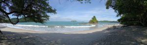 Panoramic view of a tropical beach in Manuel Antonio National Park, Costa Rica, featuring white sand, turquoise waters, lush rainforest vegetation, and a small rocky islet along the shoreline.