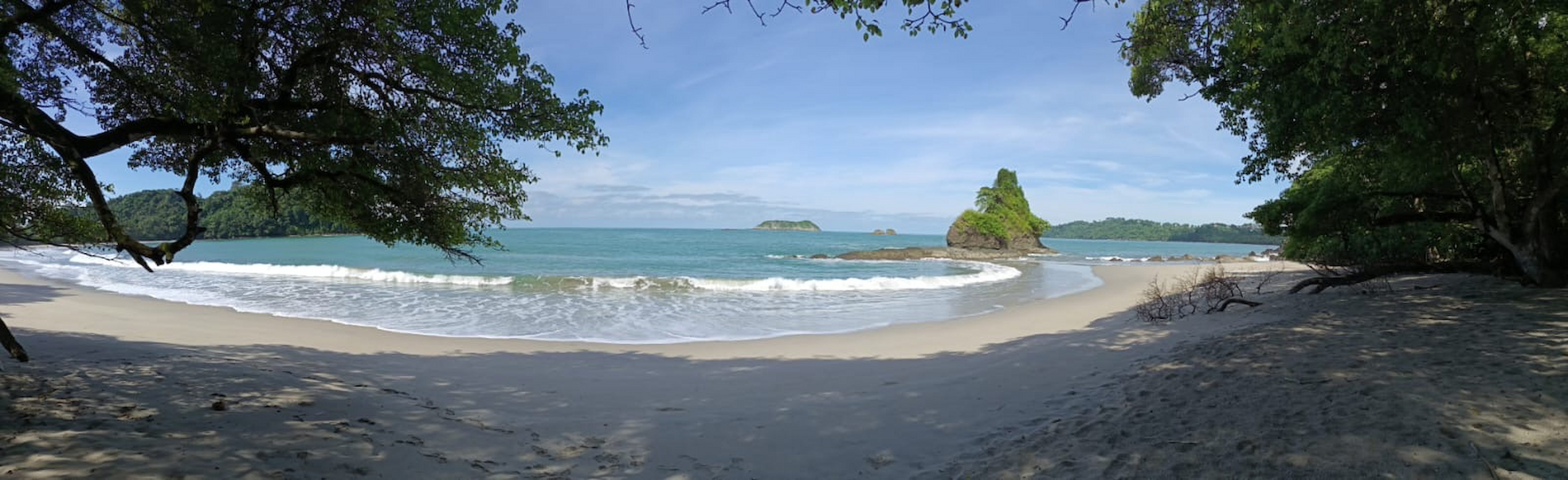 Panoramic view of a tropical beach in Manuel Antonio National Park, Costa Rica, featuring white sand, turquoise waters, lush rainforest vegetation, and a small rocky islet along the shoreline.