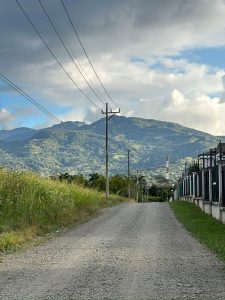 A gravel road lined with utility poles leads toward green mountains under a cloudy sky. Tall grass grows on one side, while buildings and fences line the other.