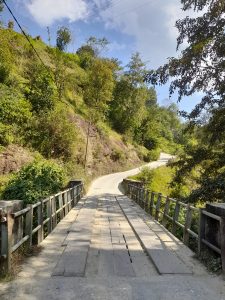 A rustic wooden bridge spans a narrow dirt road through a lush, green hillside under a partly cloudy blue sky