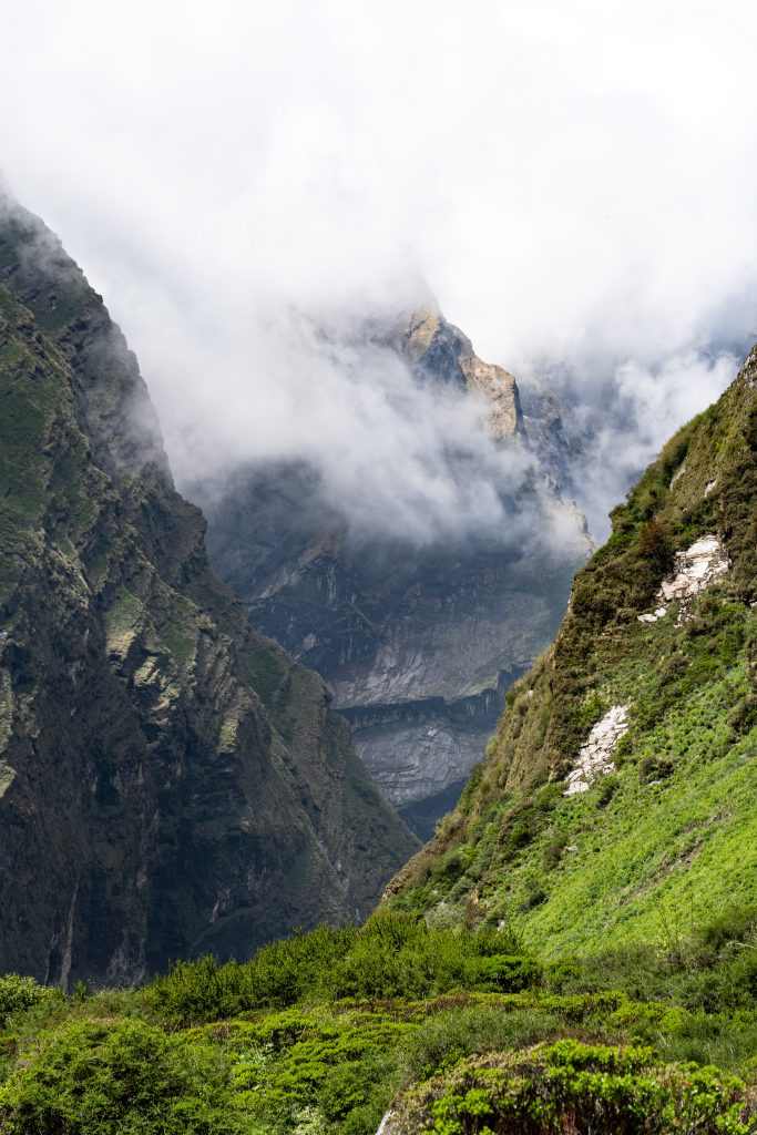 Rocky mountain gorge with a jagged peak partially shrouded in clouds, green shrubs in the foreground