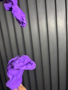 Close-up photograph of two pieces of purple modeling clay held against a dark gray corrugated metal background.