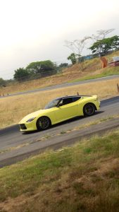 A dynamic low-angle shot of an acid-green Nissan Z sports car positioned on the asphalt track of the Autódromo de Costa Rica. Grass-covered hills, trees, and the faint outline of the track are visible beneath a cloudy sky.