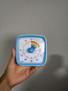 Hand holding a blue visual timer with rainbow graphics against a gray background.