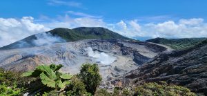 A volcanic crater with rising steam, surrounded by green mountains under a blue sky.