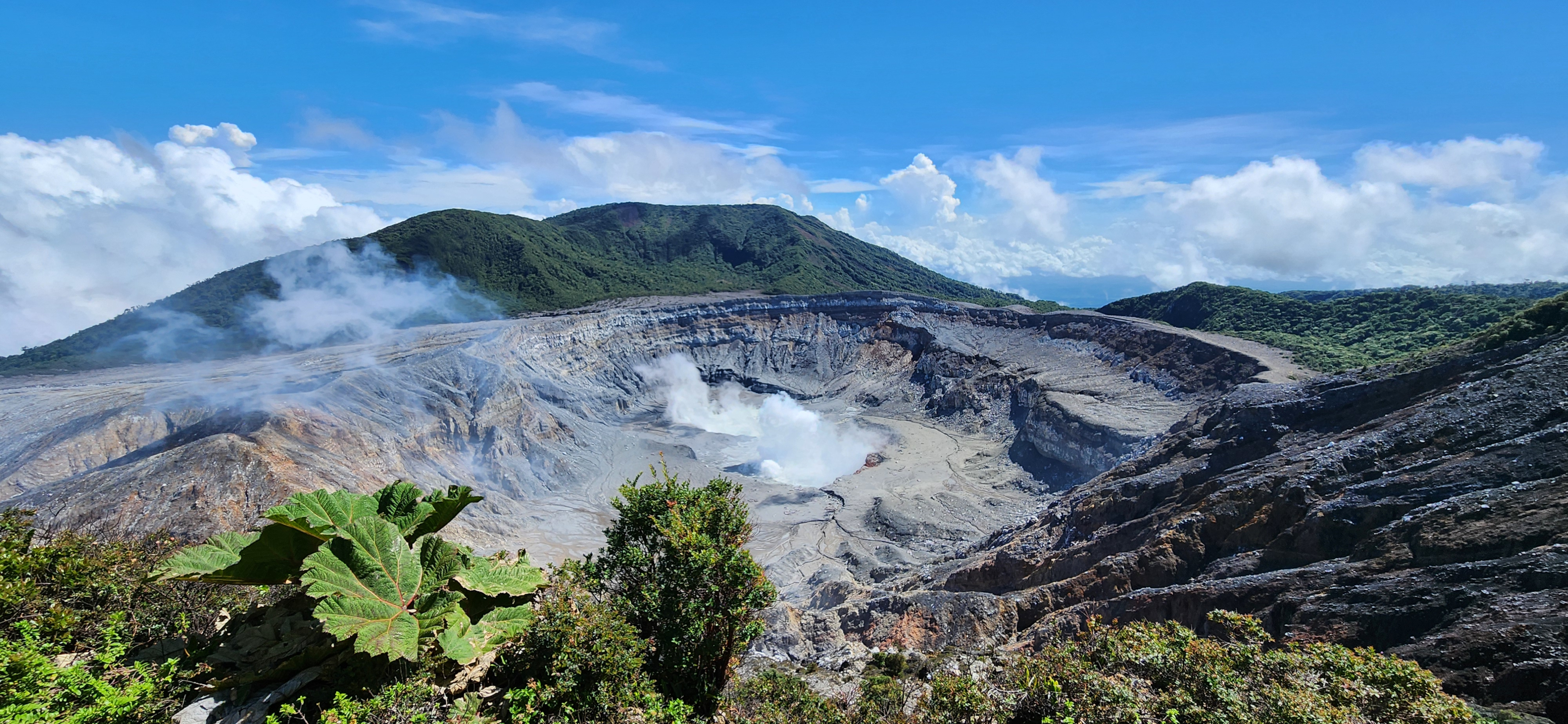A volcanic crater with rising steam, surrounded by green mountains under a blue sky.