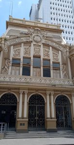 Front facade of the Correos y Telégrafos building in San José, Costa Rica, showing ornate cherub sculptures, floral relief carvings, arched entrances and wrought-iron gates.