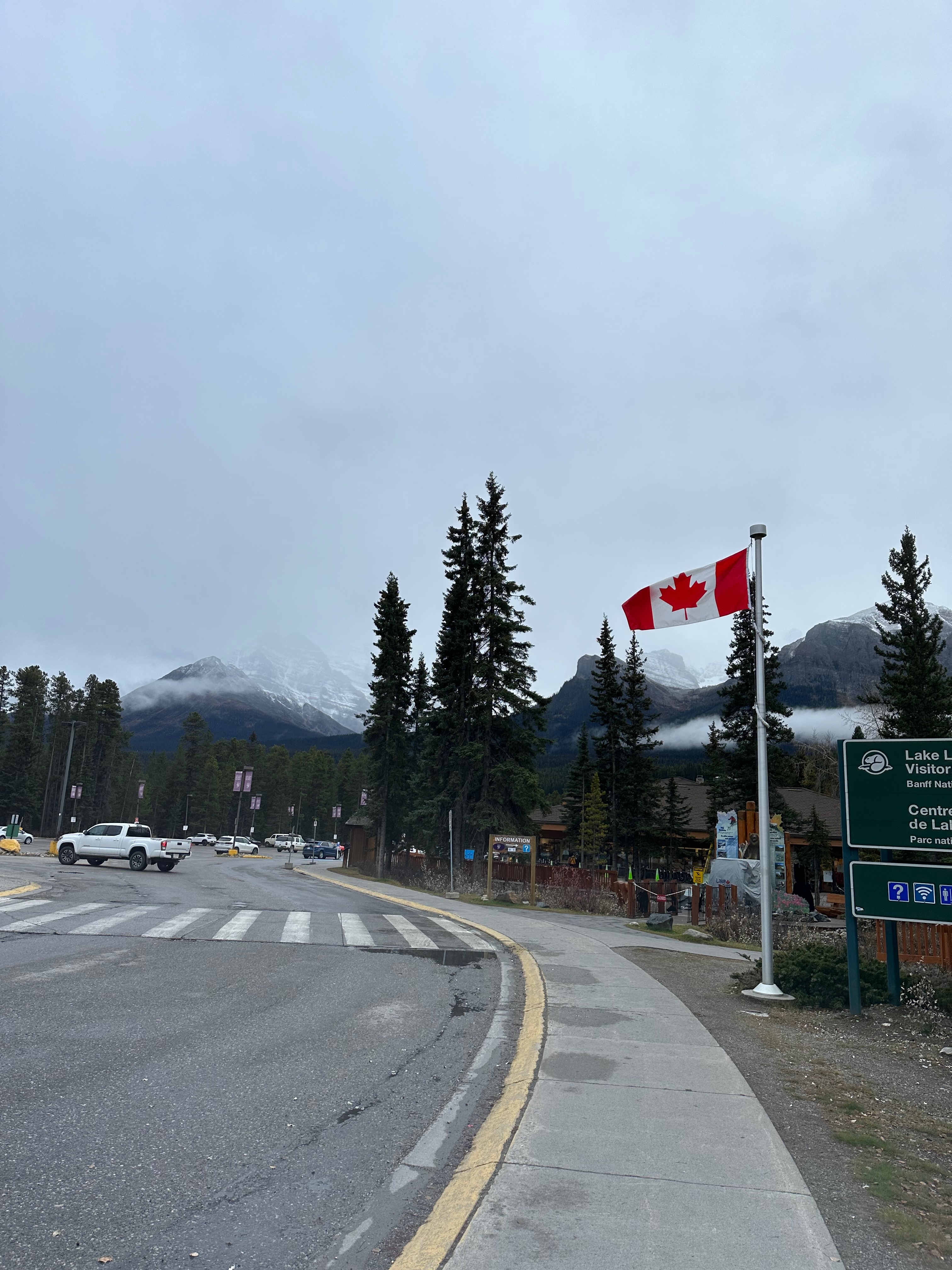 A Canadian flag waves beside a roadside visitor area, with evergreen trees, parked vehicles, and buildings at the base of misty mountain peaks. Low clouds hang over the scene under a gray sky, while a sign for the Lake Louise Visitor Centre appears on the right edge.
