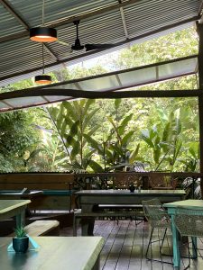 An open-air wooden restaurant with tables and chairs, surrounded by lush tropical plants.