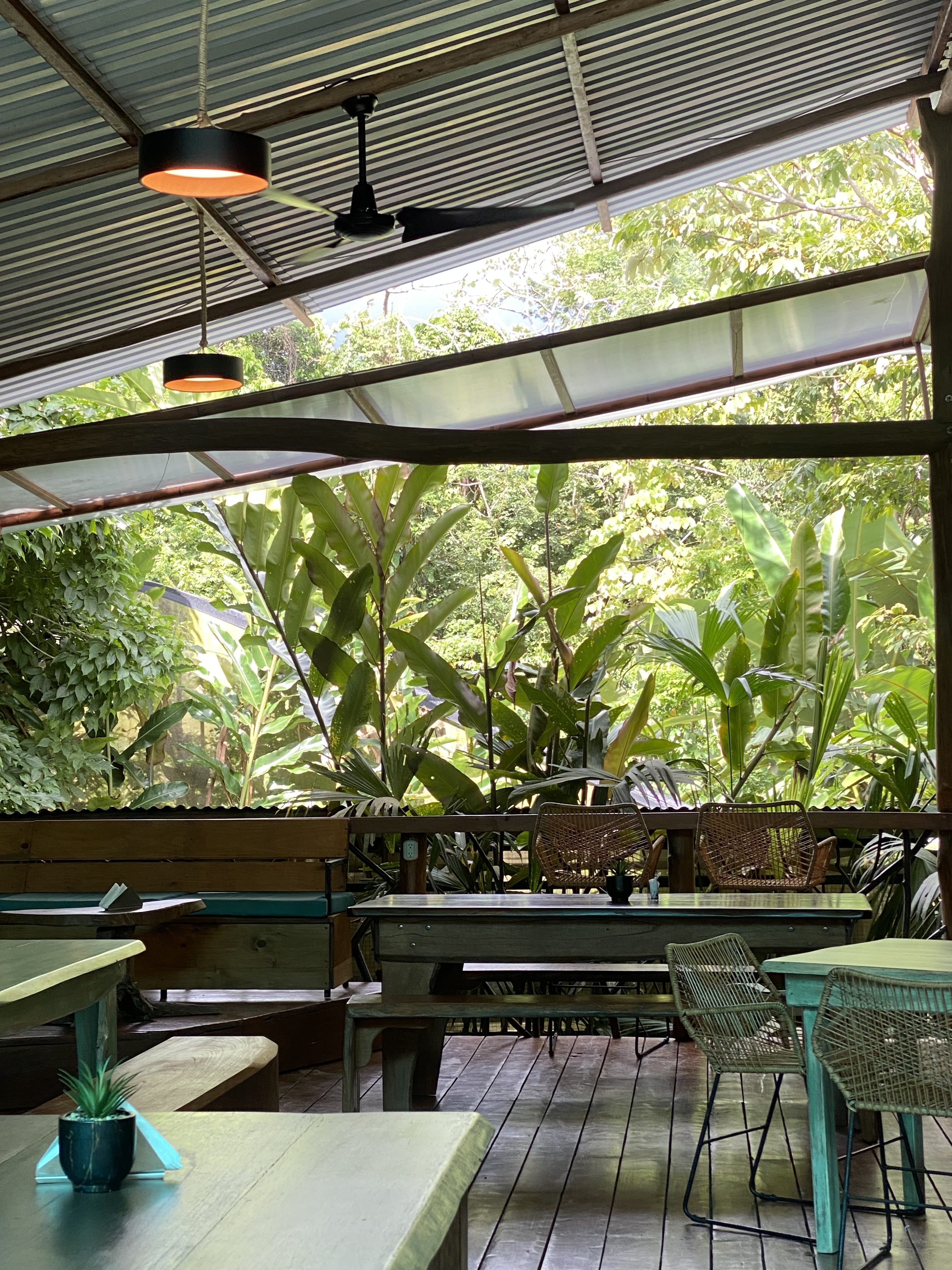 An open-air wooden restaurant with tables and chairs, surrounded by lush tropical plants.
