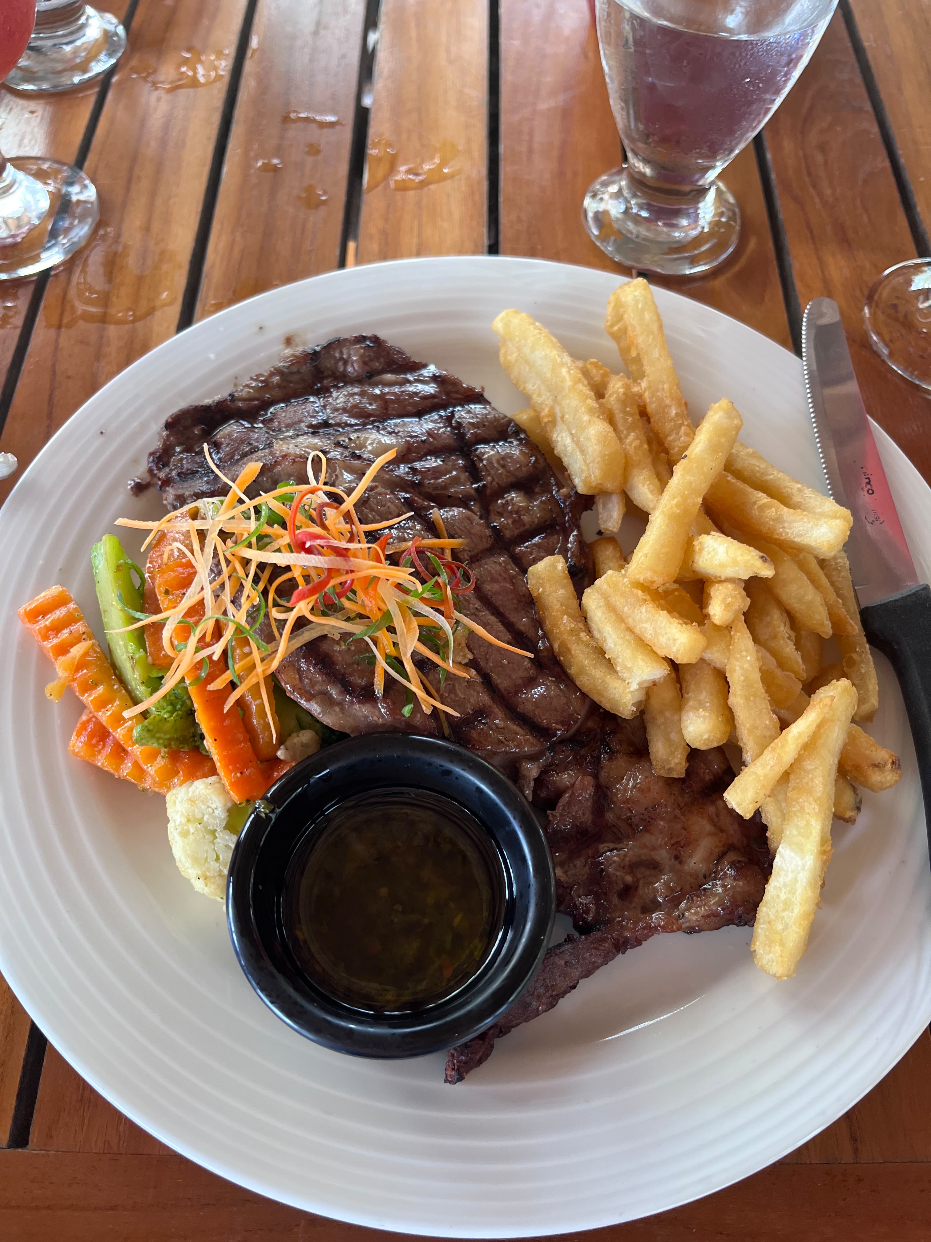 Grilled steak with vegetables and a small dish of dipping sauce on a serving plate.