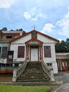 Weathered wooden chapel at the Sanatorio Durán in Costa Rica, with a curved staircase leading up to double doors beneath a cross-topped gable.
