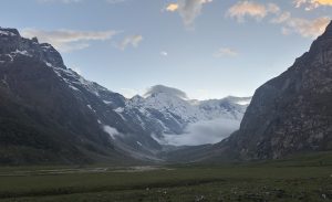 Mountain landscape at dawn with snow-capped peaks and a valley between rocky cliffs. Soft clouds hover, creating a serene and majestic atmosphere.