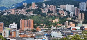 An image of a city with tall buildings and trees. The background of the city seems to be a green hilly area.