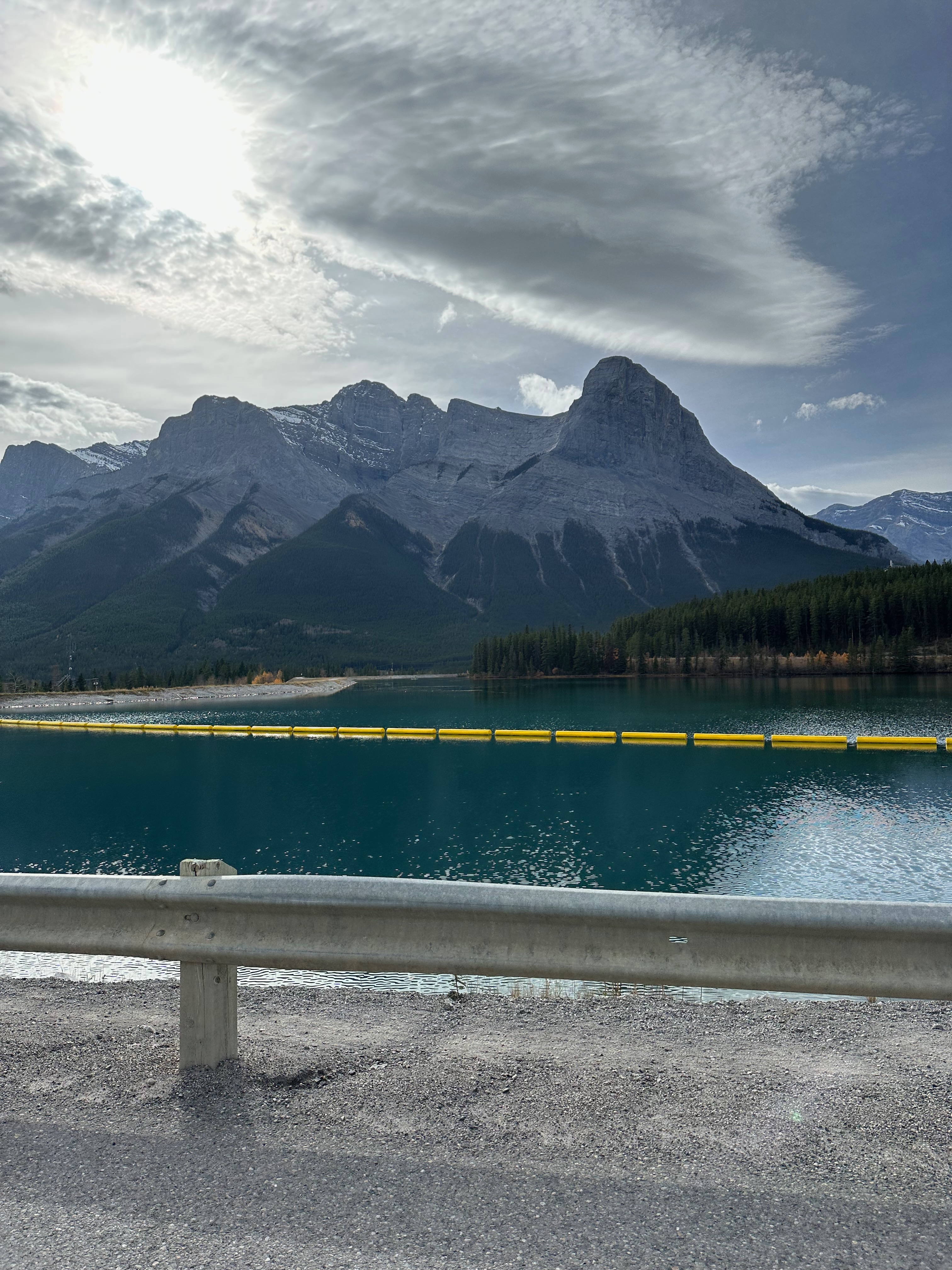 A turquoise mountain lake lies beneath towering gray peaks and dark evergreen forest. A line of bright yellow floating barriers crosses the water, while a metal roadside guardrail frames the foreground. Dramatic layered clouds stretch across the sky, casting soft shadows over the rugged landscape.